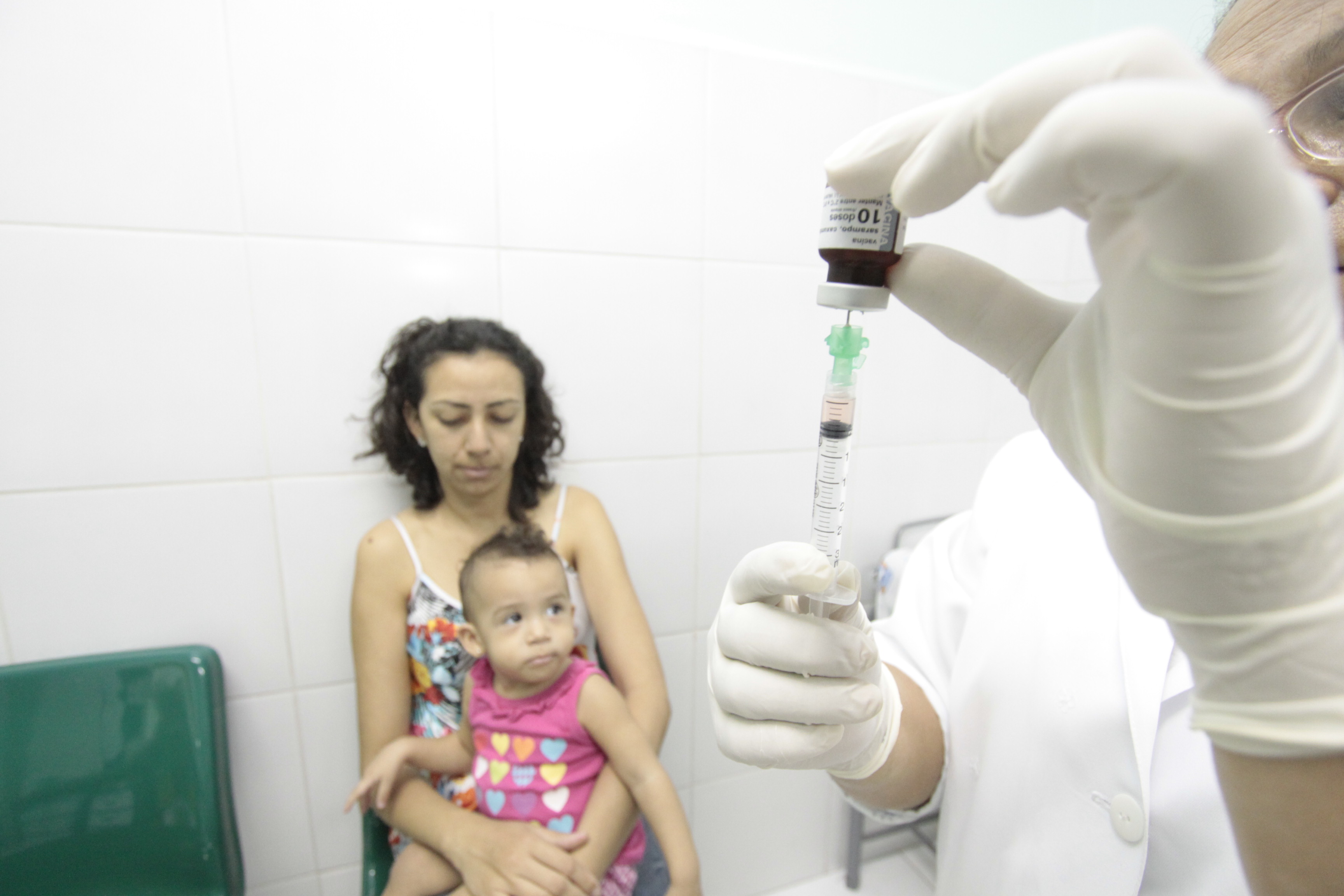 em primeiro plano, uma seringa. em segundo plano, uma mãe com uma menina de dois anos no colo